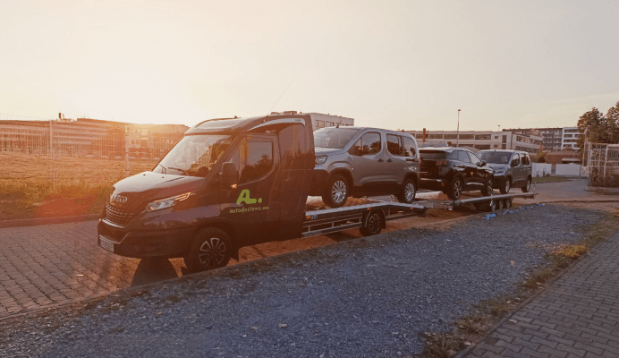 Laweta transportująca trzy pojazdy osobowe na tle zachodzącego słońca w mieście.