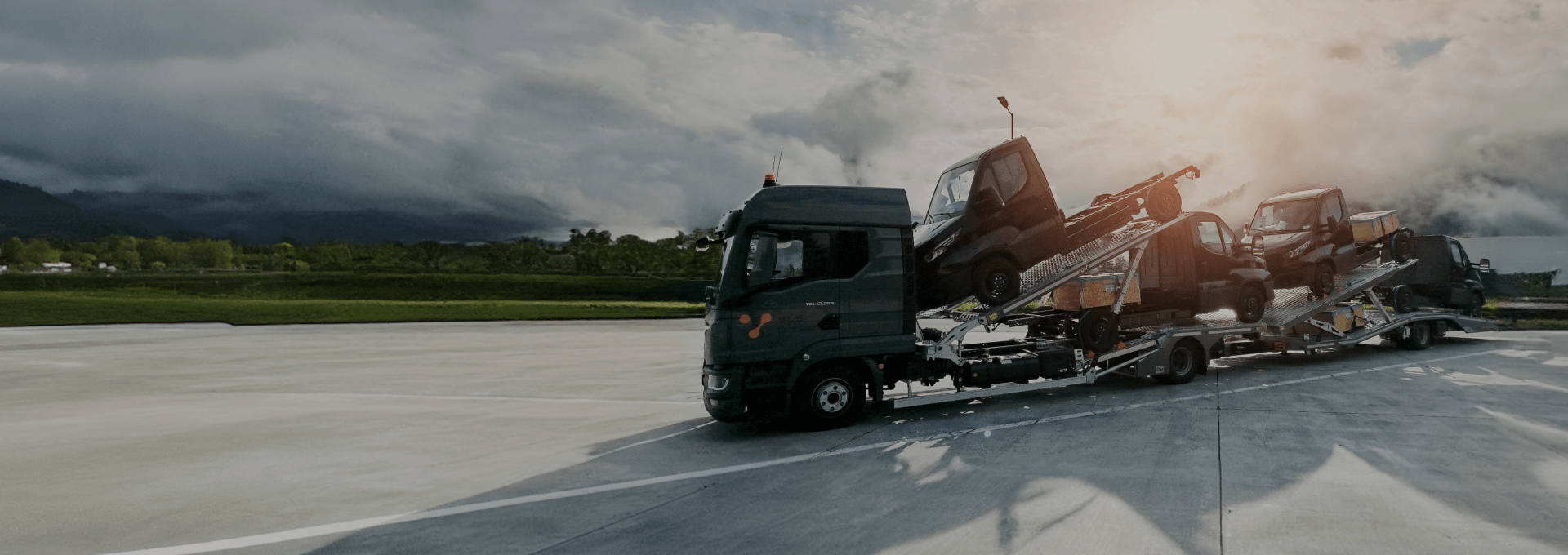 Transporter z logo VLS przewożący auta dostawcze na otwartym terenie z widokiem na zachmurzone niebo i zieleń w tle.